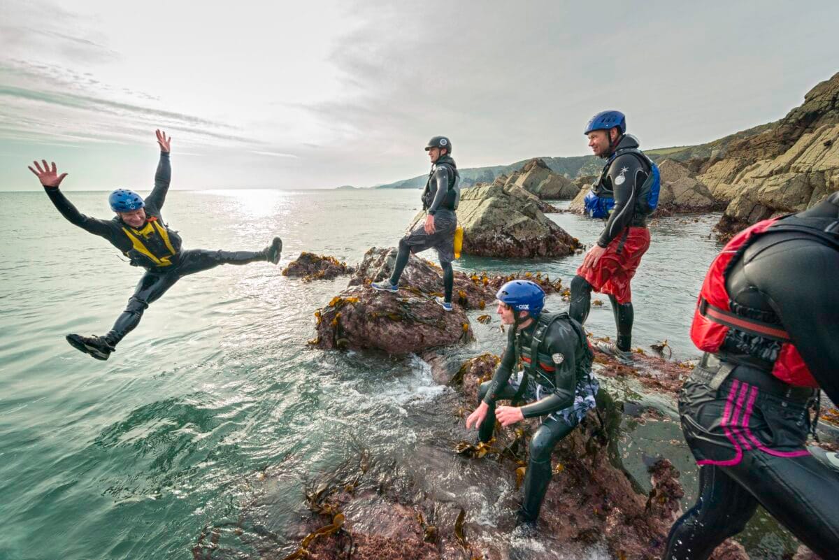 A group of people coasteering in Wales, with one person in a wetsuit and helmet jumping mid-air off a rocky cliff into the ocean while the rest of the group watches.