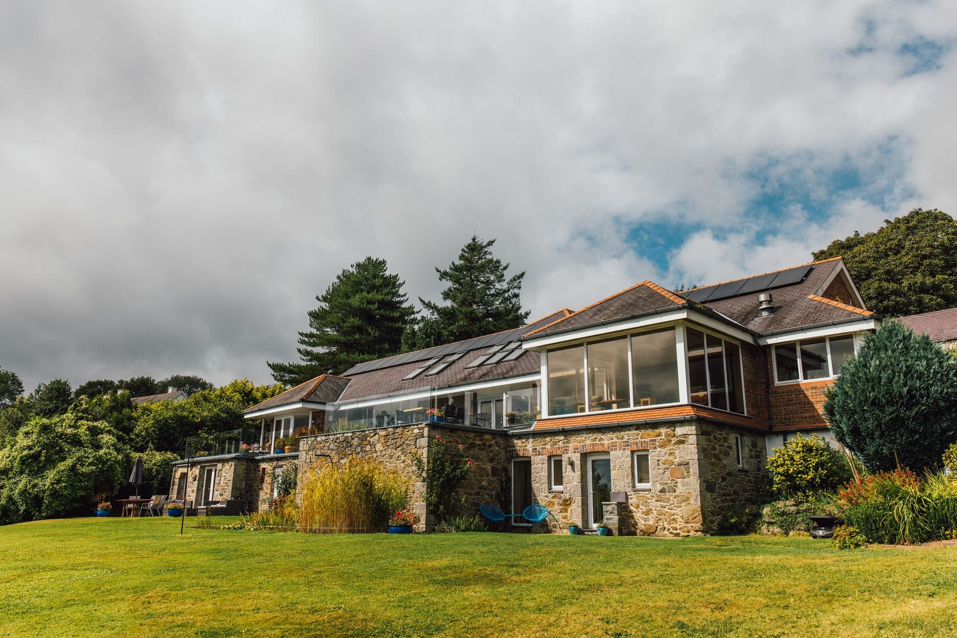 A substantial two-storey Welsh luxury holiday house set into a green hillside, built in local grey stone with red-brick window detailing, featuring a full-length glazed upper-floor sun room with glass balustrades, clay-tiled roof with integrated solar panels and rooflights, and a wide manicured lawn with ornamental grasses, planted borders and outdoor terrace furniture in the foreground, framed against a dramatic overcast Welsh sky.