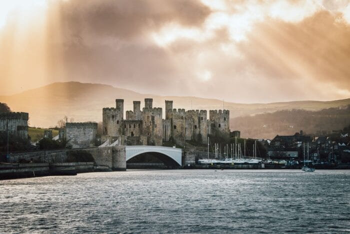 Conwy Castle, a medieval stone fortress with multiple towers, stands by a river, with boats moored nearby and a bridge in front. Sunbeams break through dramatic clouds, illuminating the landscape and distant hills.