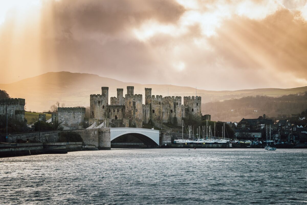 Conwy Castle in North Wales, a medieval stone fortress with multiple towers, stands by a river, with boats moored nearby and a bridge in front. Sunbeams break through dramatic clouds, illuminating the landscape and distant hills.