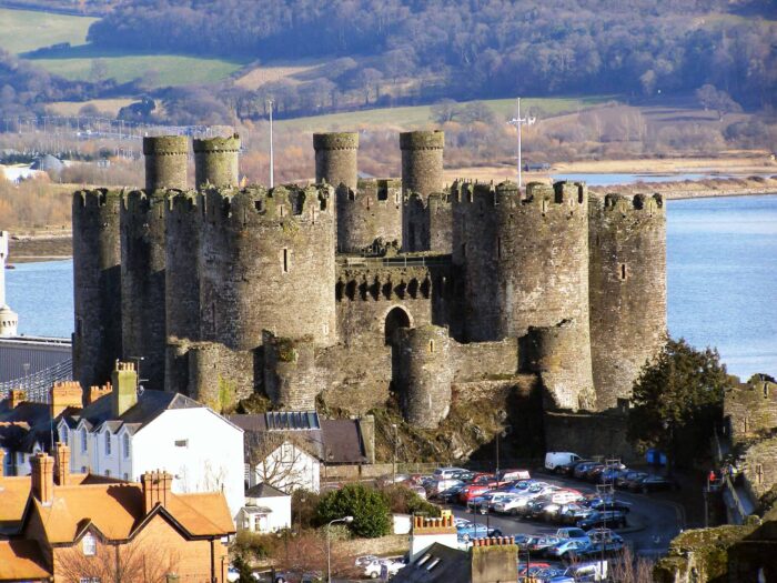 Conwy Castle, a large stone fortress with multiple round towers, sits beside a river, surrounded by parked cars and nearby houses, with green hills and trees visible in the background.