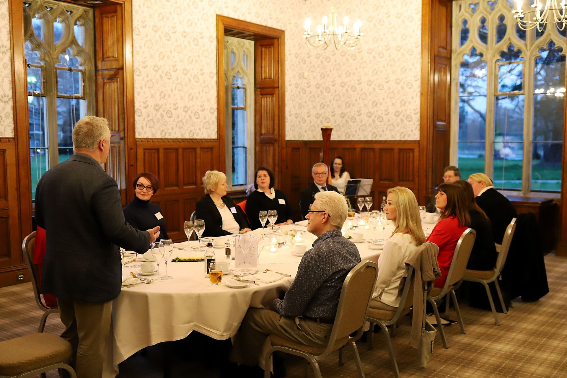 A man stands and addresses a group of professionals seated around a large, circular dining table inside a grand room at Hensol Castle, featuring rich wood paneling, patterned wallpaper, and large historic windows.