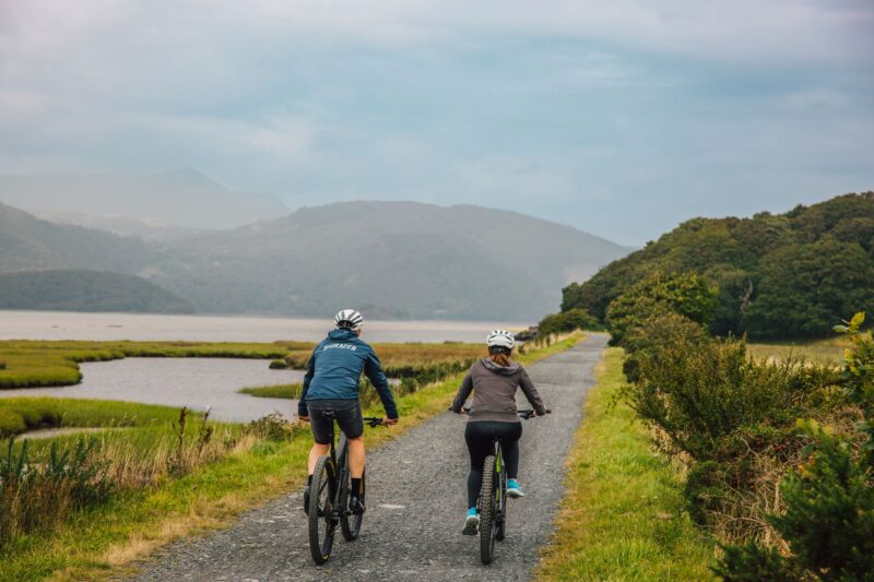 Two people wearing helmets enjoy cycling in Wales, riding bicycles on a gravel path through a scenic landscape with green hills, water, and distant mountains under a cloudy sky.