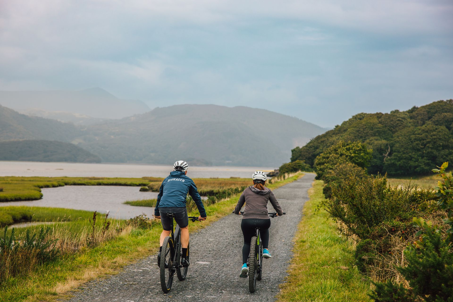 Two people wearing helmets enjoy cycling in Wales, riding bicycles on a gravel path through a scenic landscape with green hills, water, and distant mountains under a cloudy sky.