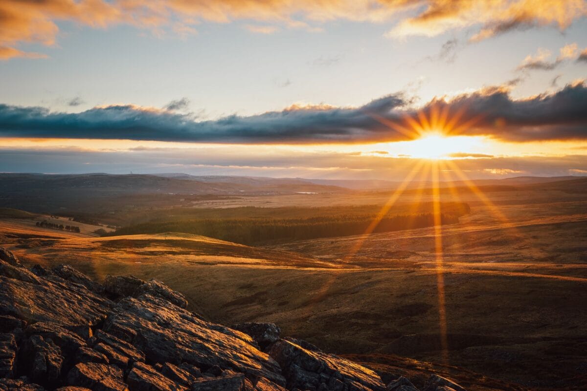 A sweeping, golden-hour landscape view of the Brecon Beacons. In the foreground, rugged, textured rocks are illuminated by the warm sunlight. Beyond them, expansive rolling hills covered in golden-brown grass and dark patches of forest stretch into the valley. A bright, dramatic sunburst shines from beneath a heavy, dark band of clouds on the right, casting striking rays of light across the entire landscape beneath a colourful sky of orange, yellow, and blue. The perfect spot to go glamping in Wales.