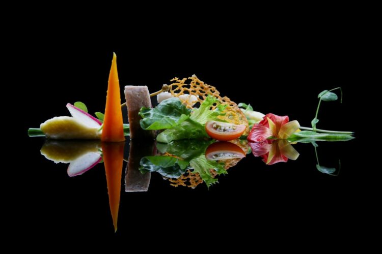 A colourful, artistic arrangement - features gourmet vegetables—a carrot spear, radish slices, greens, and edible flowers—reflected on a glossy black surface,.