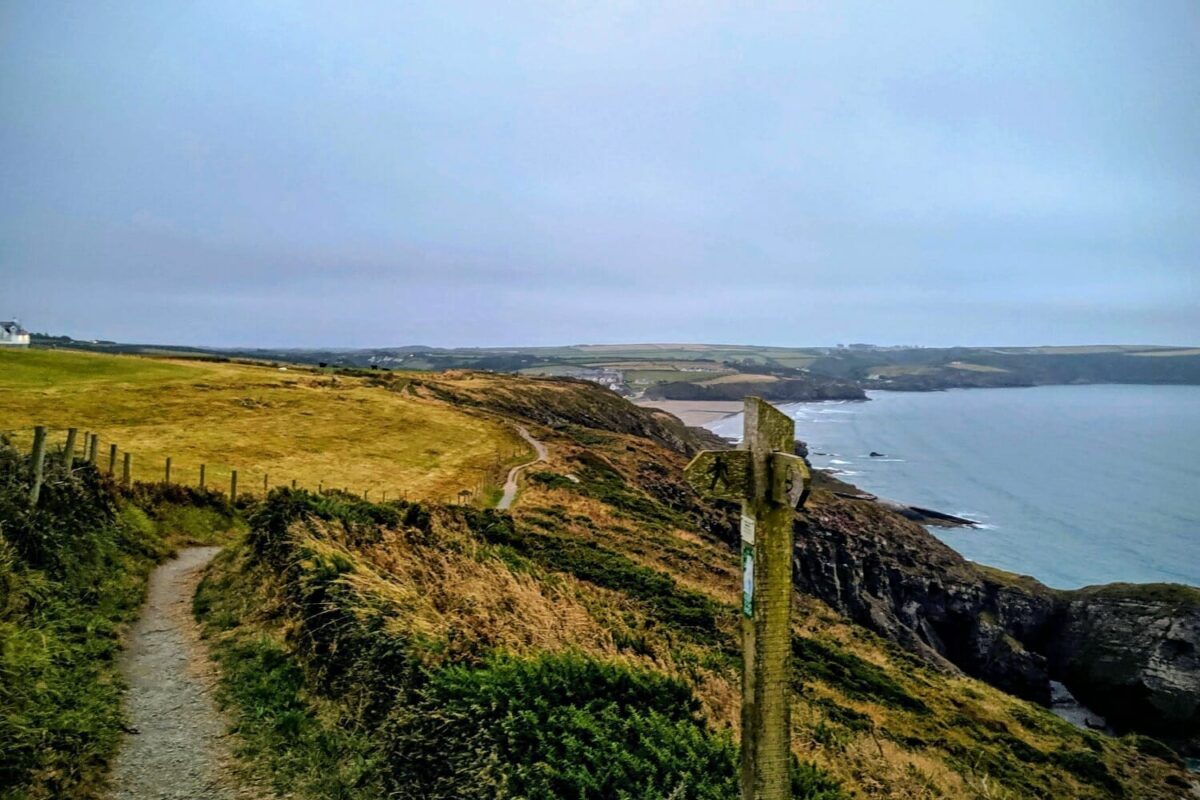 A wooden signpost marking the Pembrokeshire Coast Path as it winds along the rugged, grassy cliffs towards the sandy beach at Broad Haven
