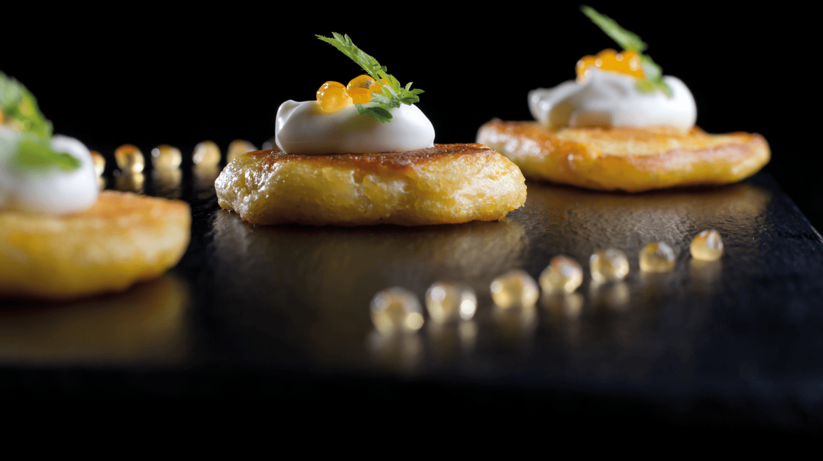 An elegant, close-up shot of gourmet appetizers featuring golden pan-fried blinis topped with rich white cream, bright orange caviar, and a delicate green garnish, beautifully plated on a dark slate surface. This fine dining dish highlights the exquisite culinary experience at Fredricks at Machynys, one of the standout restaurants in Llanelli.