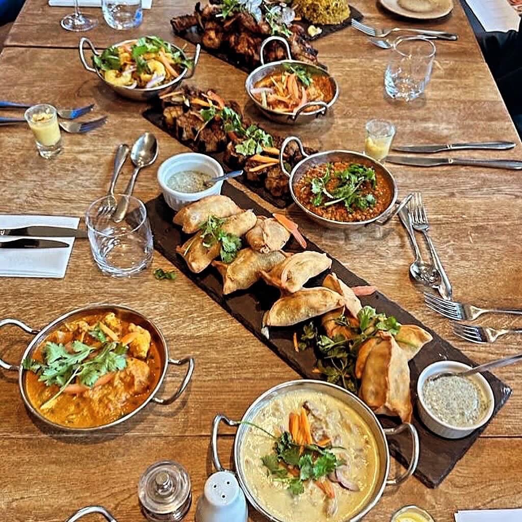 A large wooden dining table filled with a vibrant feast of Indian cuisine, featuring rich curries garnished with fresh herbs in metal serving bowls, golden samosas on a long dark slate platter, and grilled meats. 