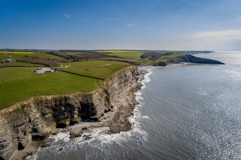 Aerial view of rocky cliffs and a rugged coastline along the Glamorgan Heritage Coast coastal walk in South Wales, with green fields above meeting the calm sea under a clear blue sky.