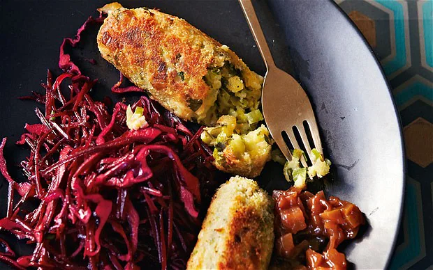 A plate with two golden-brown, breaded vegetarian Glamorgan sausages, one cut open with a fork, served with shredded red cabbage and a portion of chutneys.