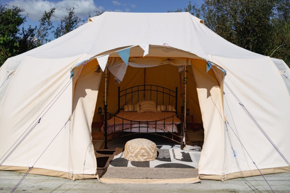 A bright, white canvas bell tent pitched on a wooden deck, with its front flaps tied back to reveal a cozy interior. Above the entrance hangs a string of pastel-colored bunting. Inside, the tent is furnished with a black metal-framed double bed, layered with patterned pillows and throws. A woven pouf sits near the entrance on top of layered, geometric-patterned rugs. The tent is set against a backdrop of green trees and a blue sky with scattered clouds.