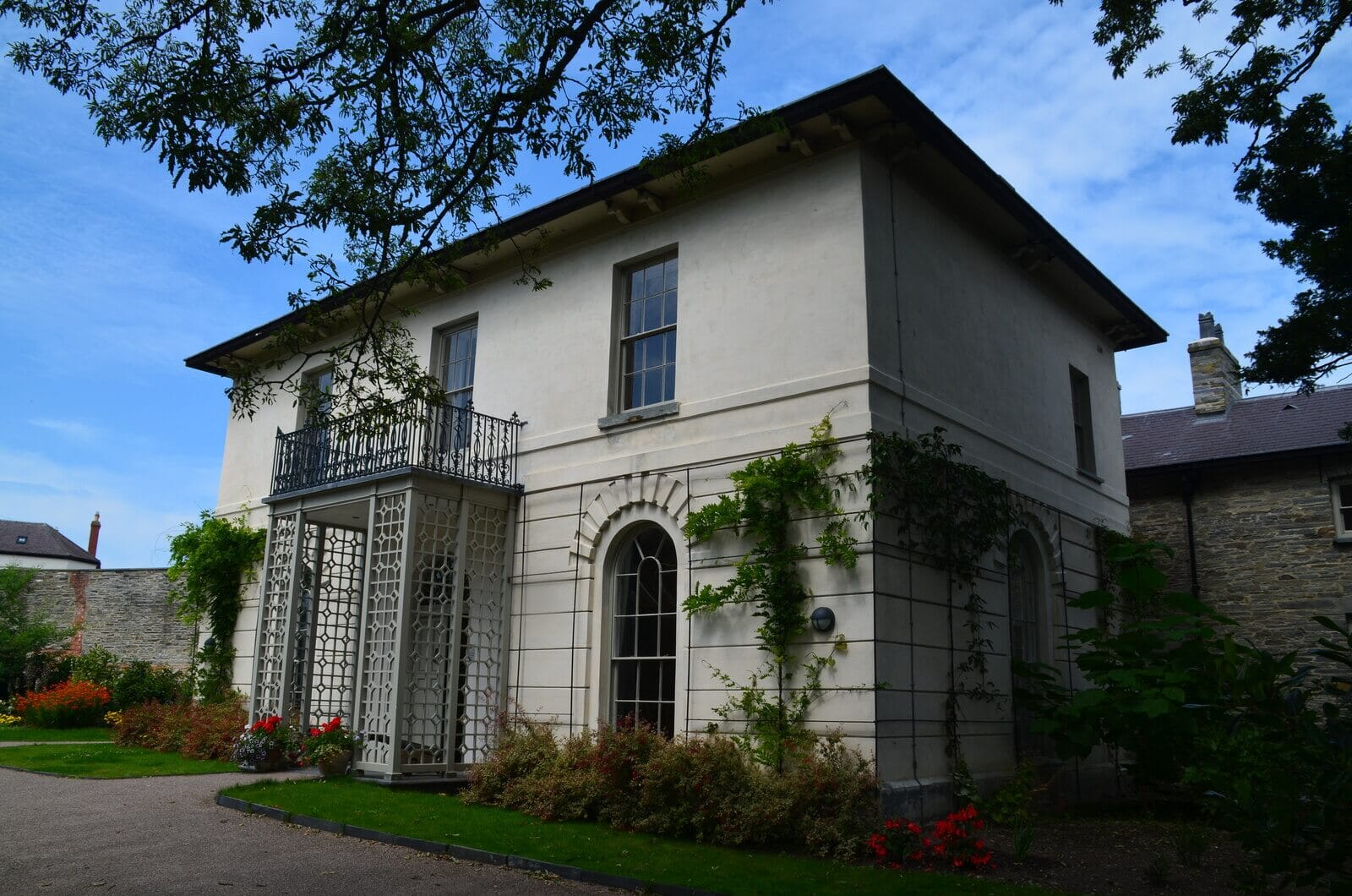 Castle Green House at Cardigan Castle, a cream-coloured two-storey Georgian house with a wrought-iron balcony above a lattice porch, an arched window and climbing plants, set in a garden with flower beds.