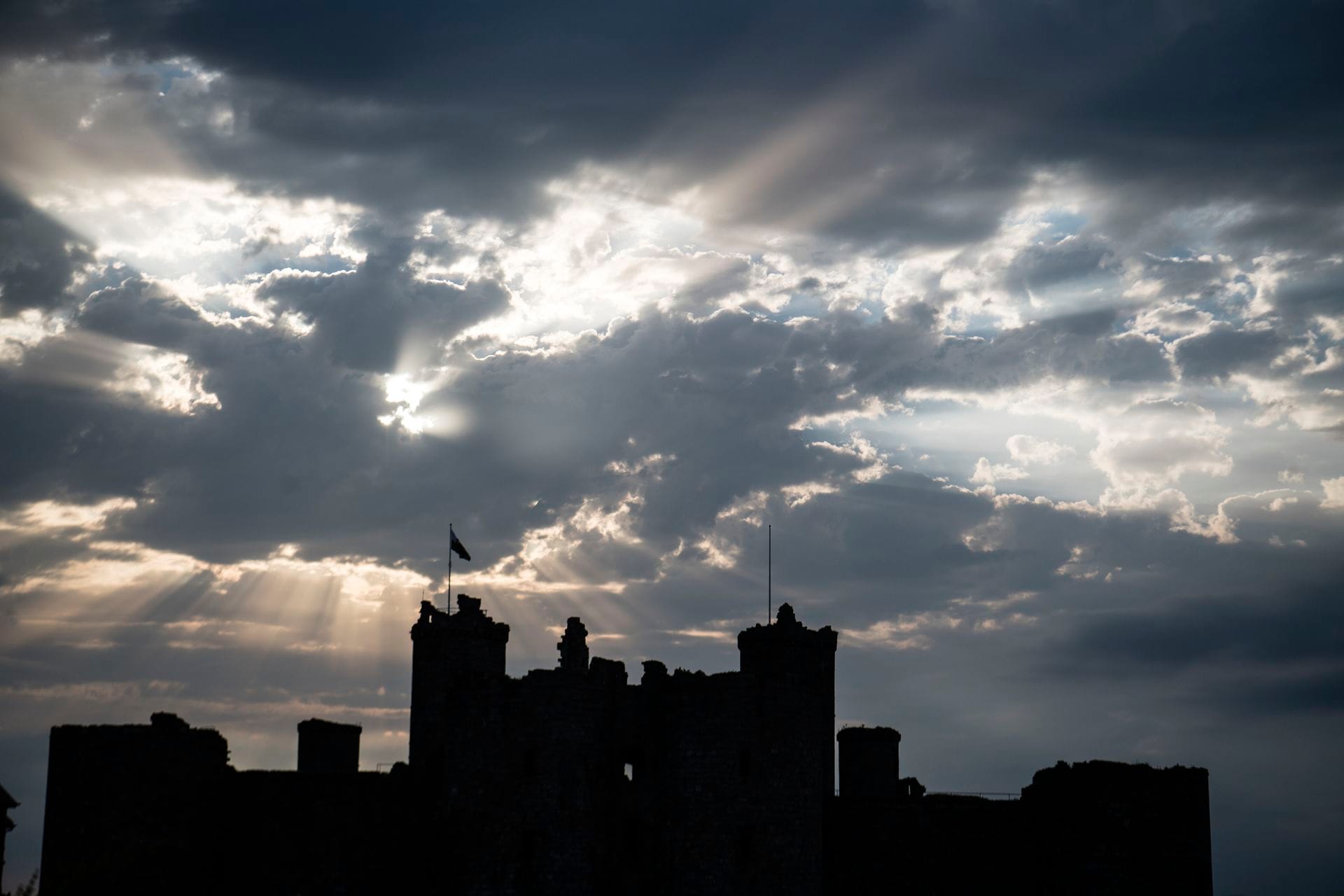Harlech Castle's towers silhouetted in black against a dramatic sky of golden sunbeams breaking through storm clouds at dusk, with two flags visible on the battlements