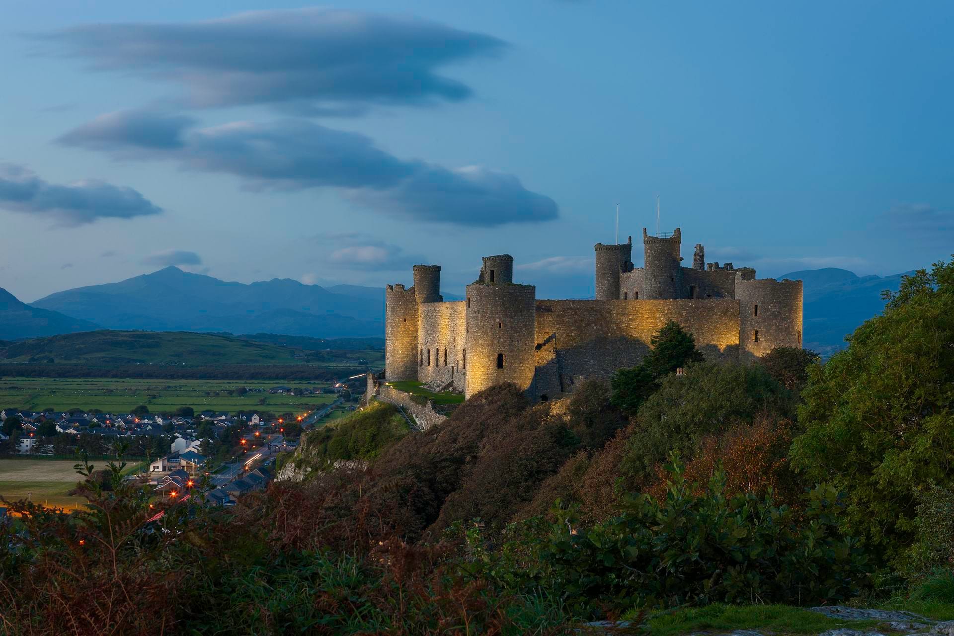 A twilight view of Harlech Castle perched high on a dark, wooded rocky crag. The castle's massive stone walls and towers are illuminated by warm golden floodlights, standing out against a deep blue evening sky. Distant mountains rise in the background, and the lights of a small coastal town twinkle on the flat plains below to the left.