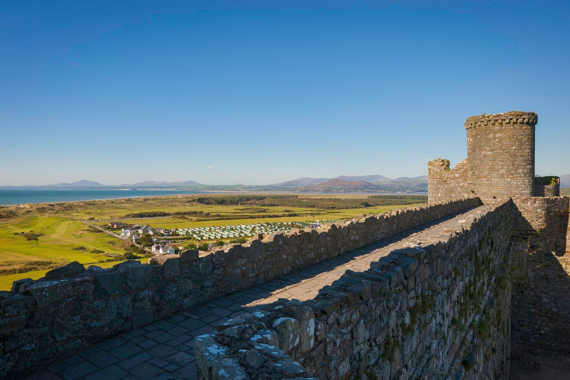 A view from Harlech Castle's wall walk looking out over green coastal grassland and sand dunes toward the sea, with distant mountains visible across the bay under a clear blue sky
