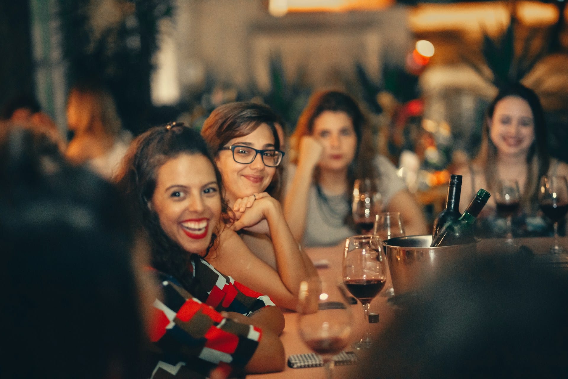 A group of women smiling and socializing around a table in a dimly lit, atmospheric bar. In the foreground, two friends smile warmly at the camera, with wine glasses and a bottle cooler resting on the table in front of them.