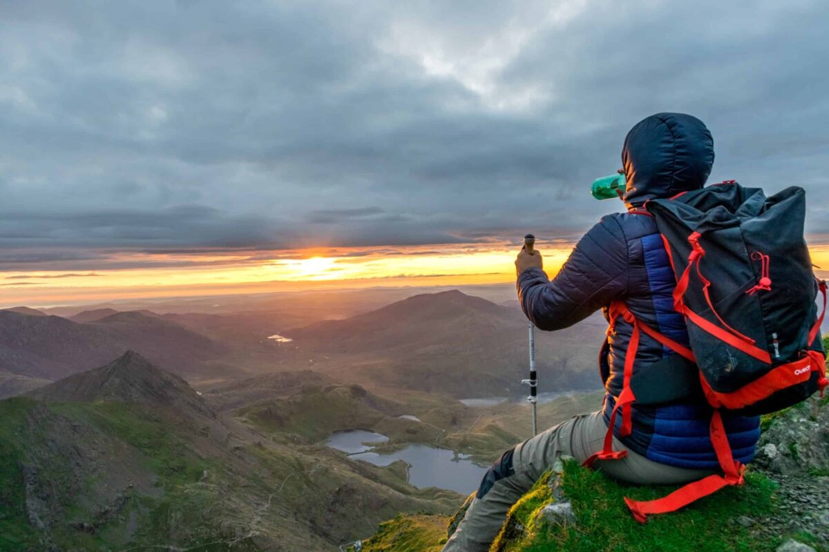 A hiker in a hooded jacket, holding a trekking pole, sits on a grassy mountain peak during mountain walks in Snowdonia, sipping from a bottle whilst watching the sun set over mountains and lakes below.