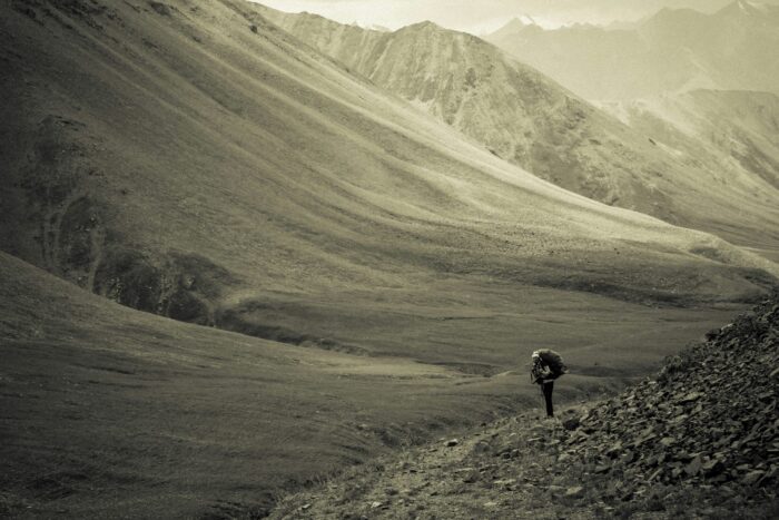 A lone hiker with a rucksack walks along a narrow path through a vast, rugged mountain valley—an inspiring scene reminiscent of day walks in Wales, where expansive, barren landscapes and distant peaks unfold beneath a cloudy sky.