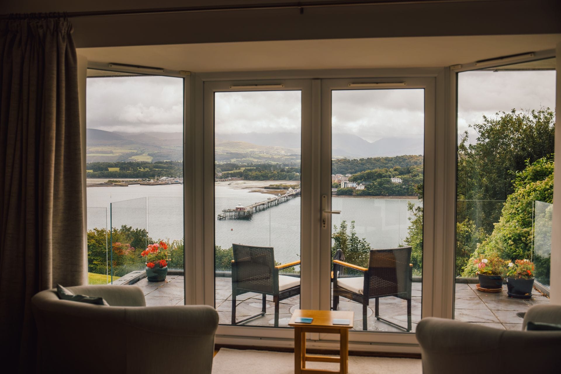 View from inside a holiday cottage looking through large glass patio doors onto a balcony with two chairs, overlooking a calm estuary with a long wooden pier stretching into the water, surrounded by green trees and distant mountains under a cloudy sky.