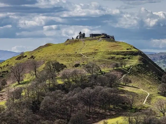 The Horseshoe Pass cycling route. A hilltop with scattered trees leads to the ruins of a stone castle at the summit, under a cloudy sky with patches of sunlight. Winding paths cross the grassy hillside,