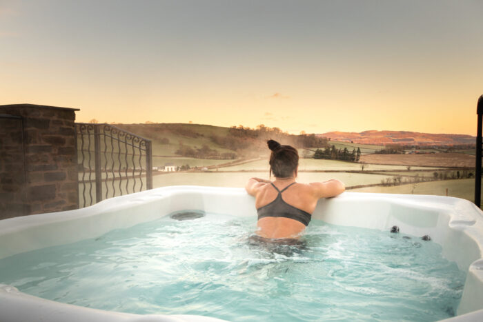 A woman in a swimming costume relaxes in a hot tub at a cottage with a hot tub in Wales, facing a scenic view of rolling hills and a river at sunset, with steam rising from the water.