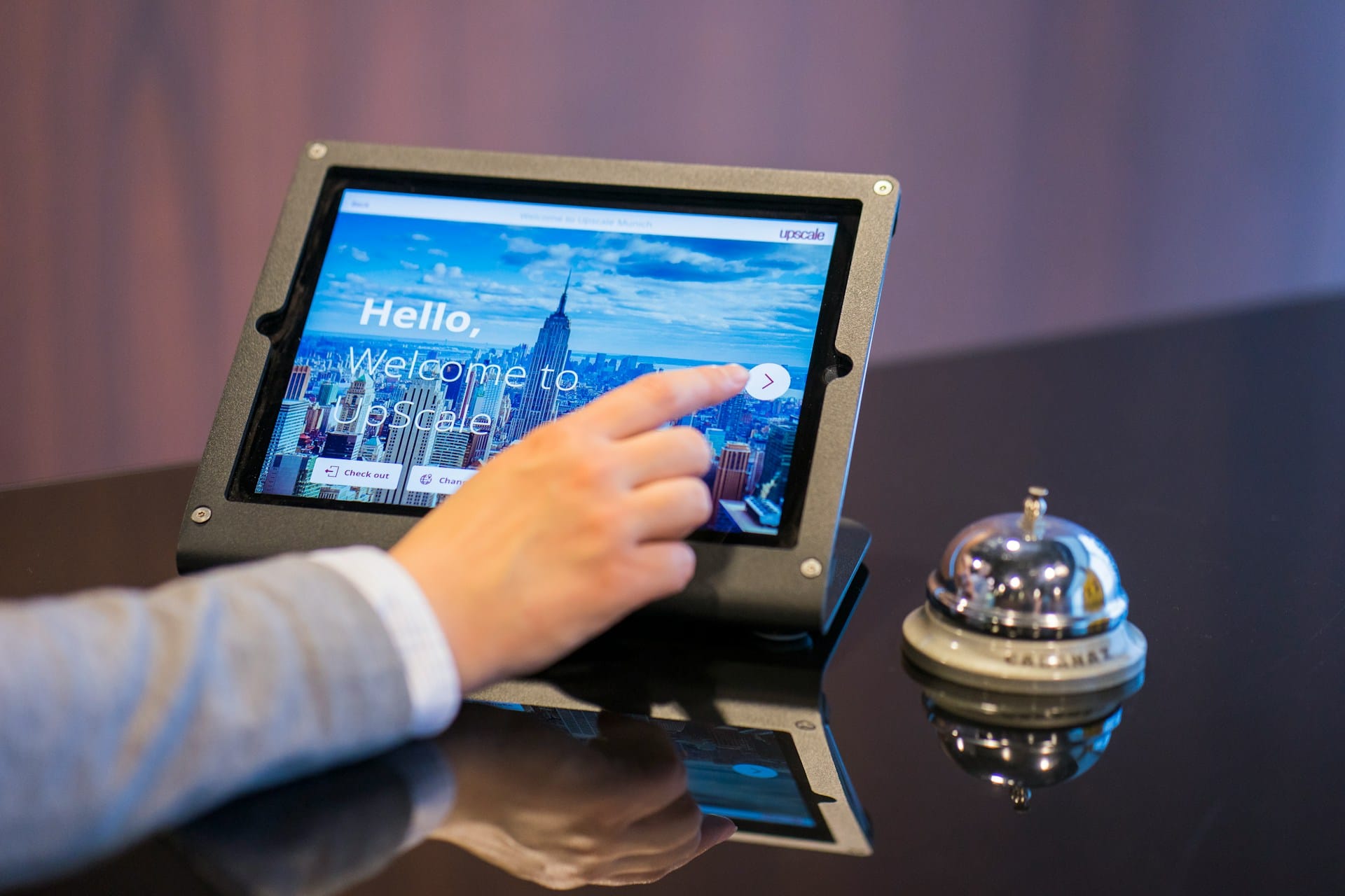 A hand touching a welcome screen on a tablet mounted at a reception desk, with a silver service bell beside it.