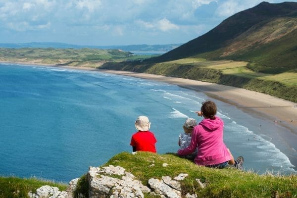 A mother and her two young children sitting on a grassy cliff top, looking out over the expansive sandy beach and blue waters of Rhossili Bay. The mother, wearing a pink hoodie, is pointing down towards the beautiful curving coastline, which is framed by green rolling hills under a partly cloudy blue sky.