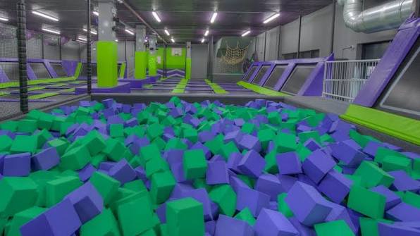 A brightly lit indoor trampoline park featuring a large foam pit filled with green and purple square blocks in the foreground, leading out to a sprawling room of interconnected flat and angled wall trampolines framed in matching purple and green padding.