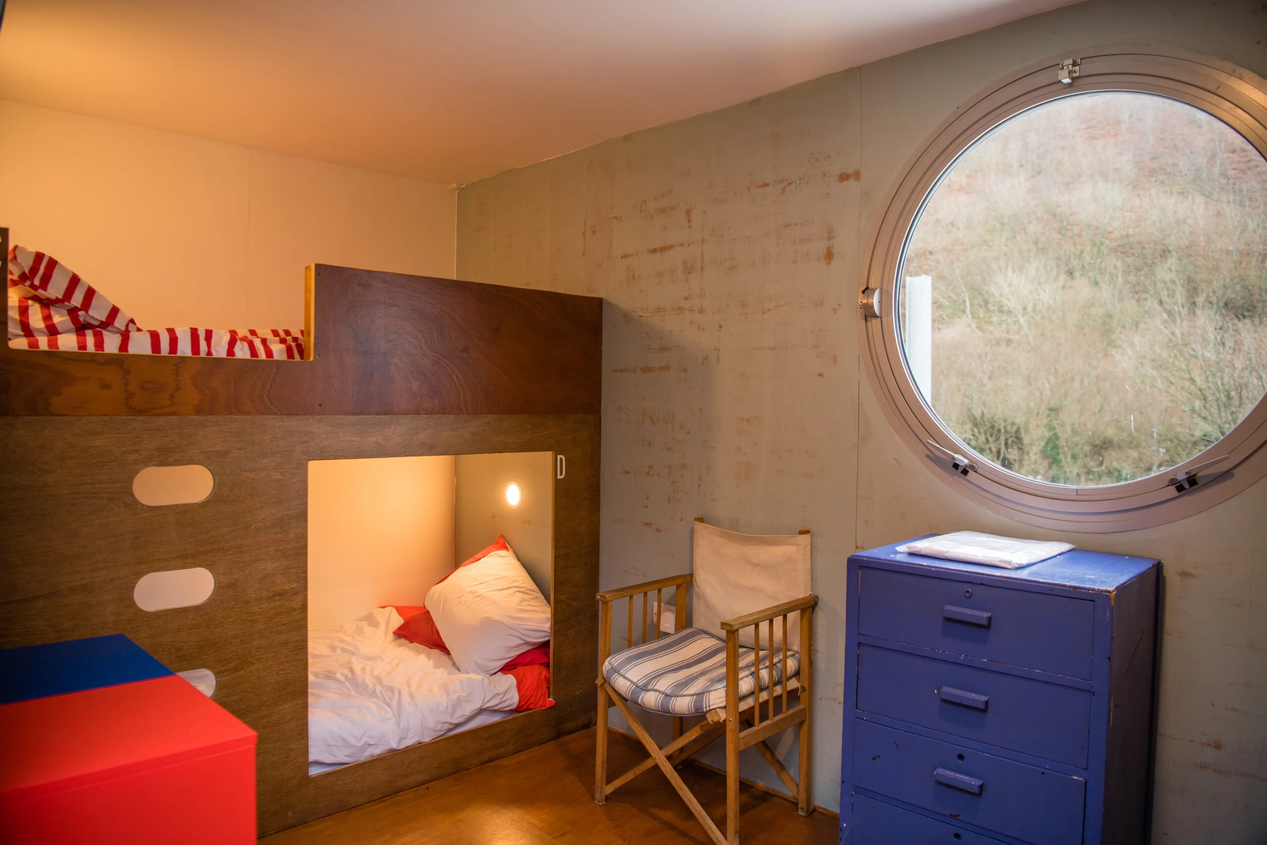 The interior of a sleeping cabin at Tŷ Cwch Boathouse. Built-in wooden pod-style bunk beds with cozy bedding and warm reading lights sit against the wall. A large circular porthole window looks out onto a steep green hillside, with a blue wooden chest of drawers and a folding chair completing the repurposed shipping container space.
