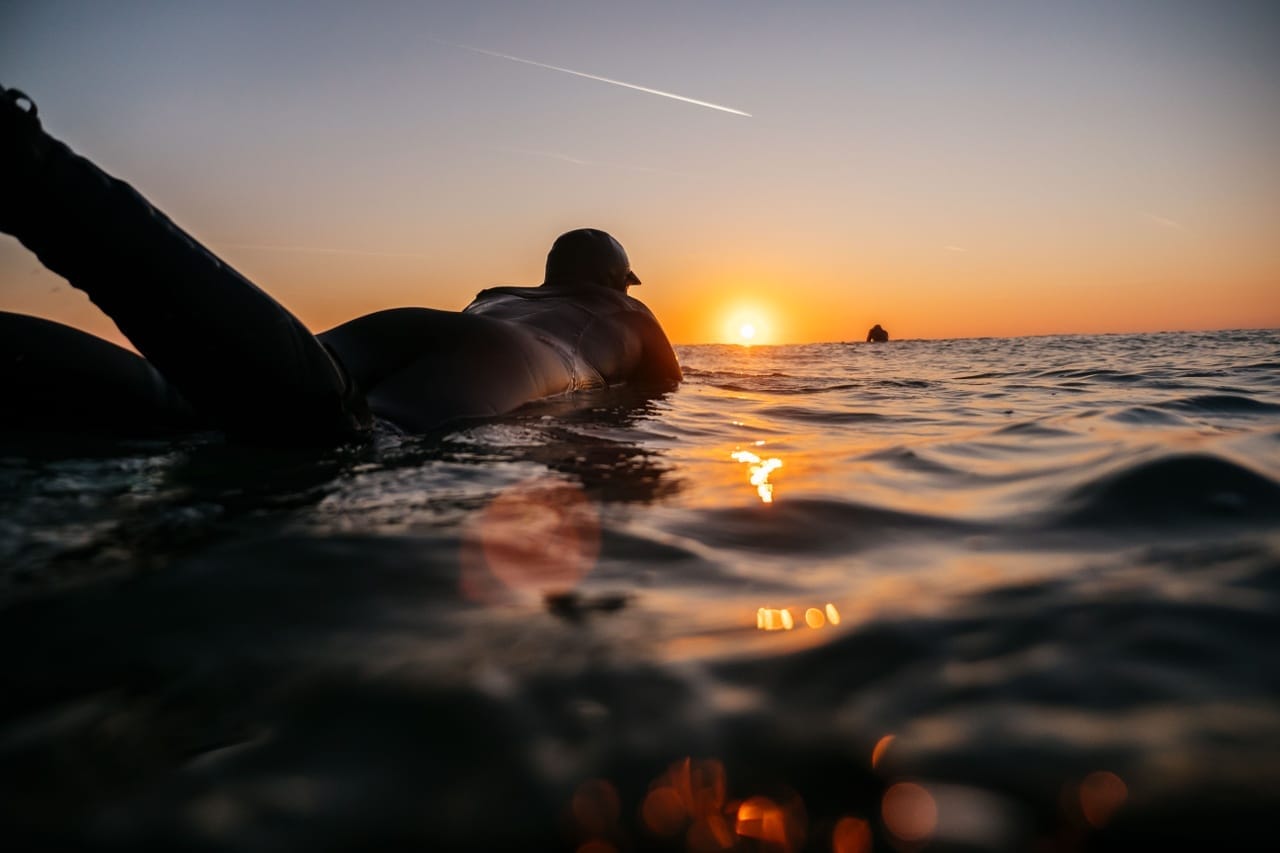 A low-angle view of a person in a wetsuit resting in the ocean water, silhouetted against a vibrant golden sunset that reflects brightly off the rippling waves.