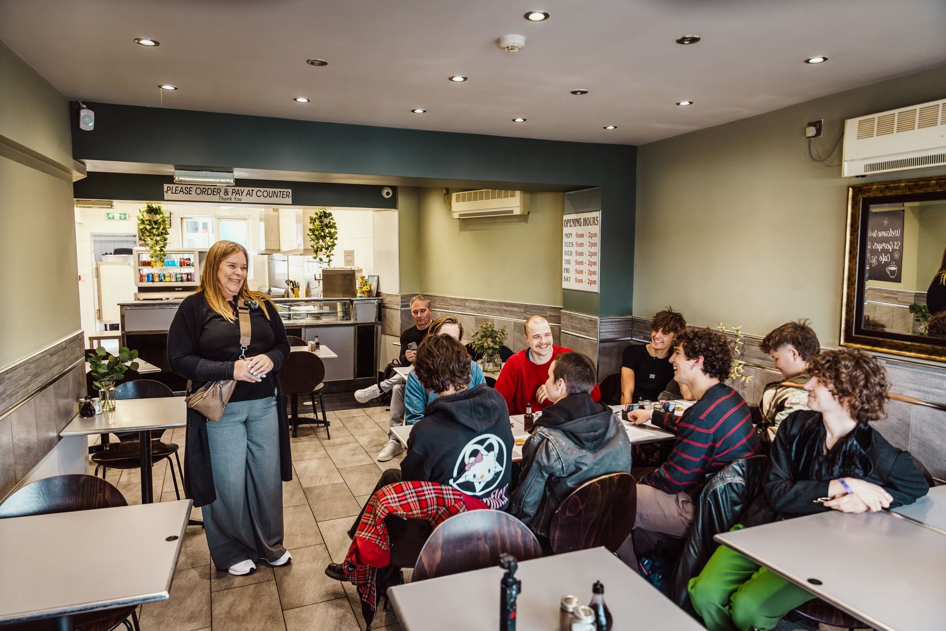 A female tour guide stands and speaks to a seated group of smiling young adults gathered around tables inside a traditional Welsh fish and chip shop, with the service counter visible in the background.