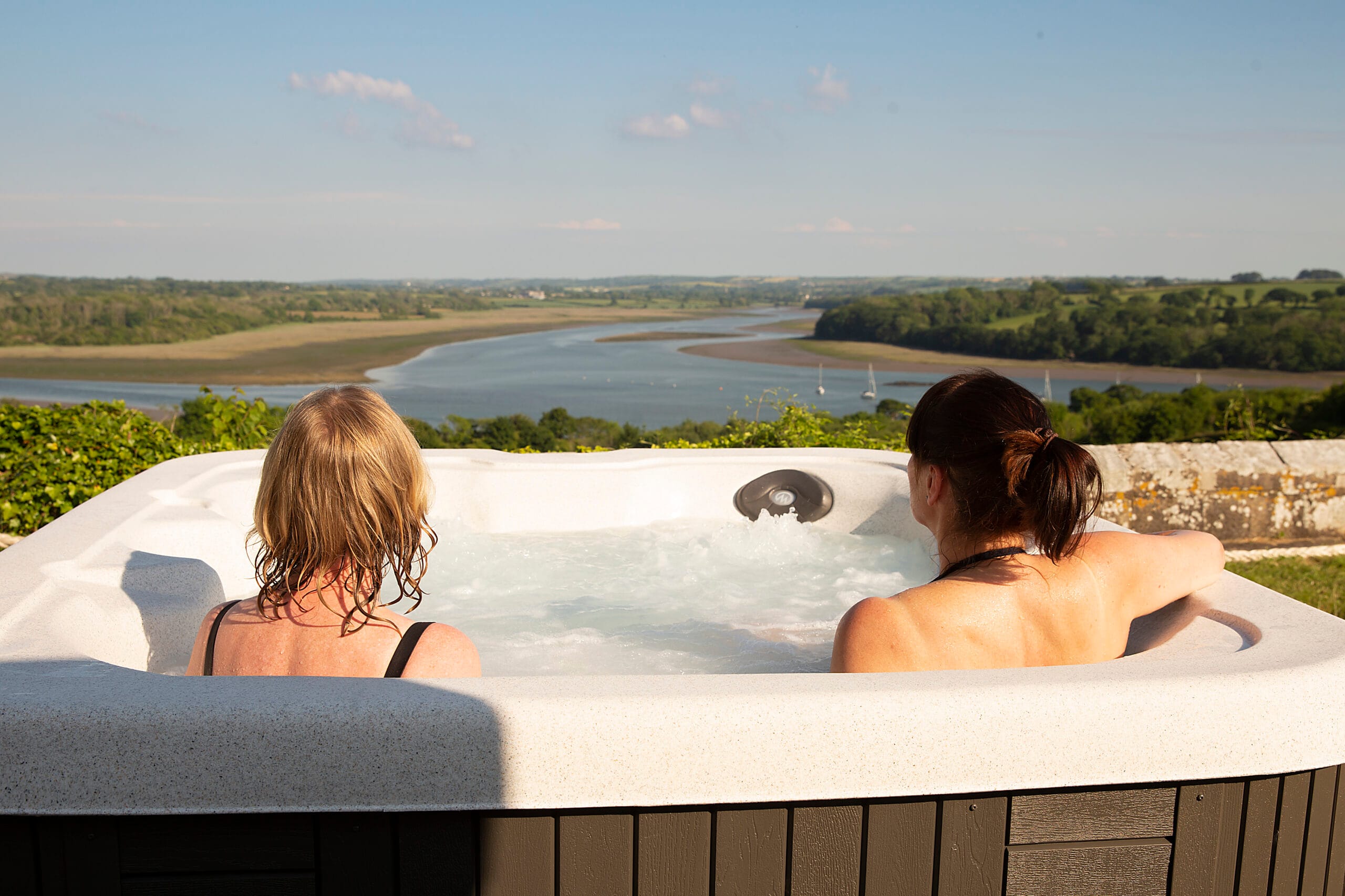 Two women relaxing in an outdoor hot tub, looking out over a wide, scenic Welsh estuary surrounded by rolling green hills under a bright blue sky.