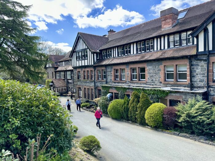 A group of people walk along a road beside the historic Lake Vyrnwy Hotel in Llanwddyn, featuring Tudor-style woodwork and surrounded by greenery and bushes under a partly cloudy Montgomeryshire sky.