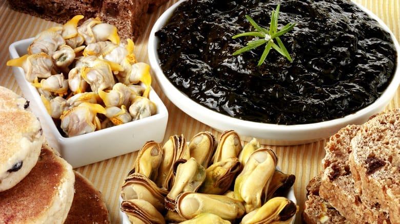 A close-up of a traditional Welsh meal featuring laverbread in a white bowl, surrounded by baked bread, Welsh cakes, and dishes of cooked cockles and mussels