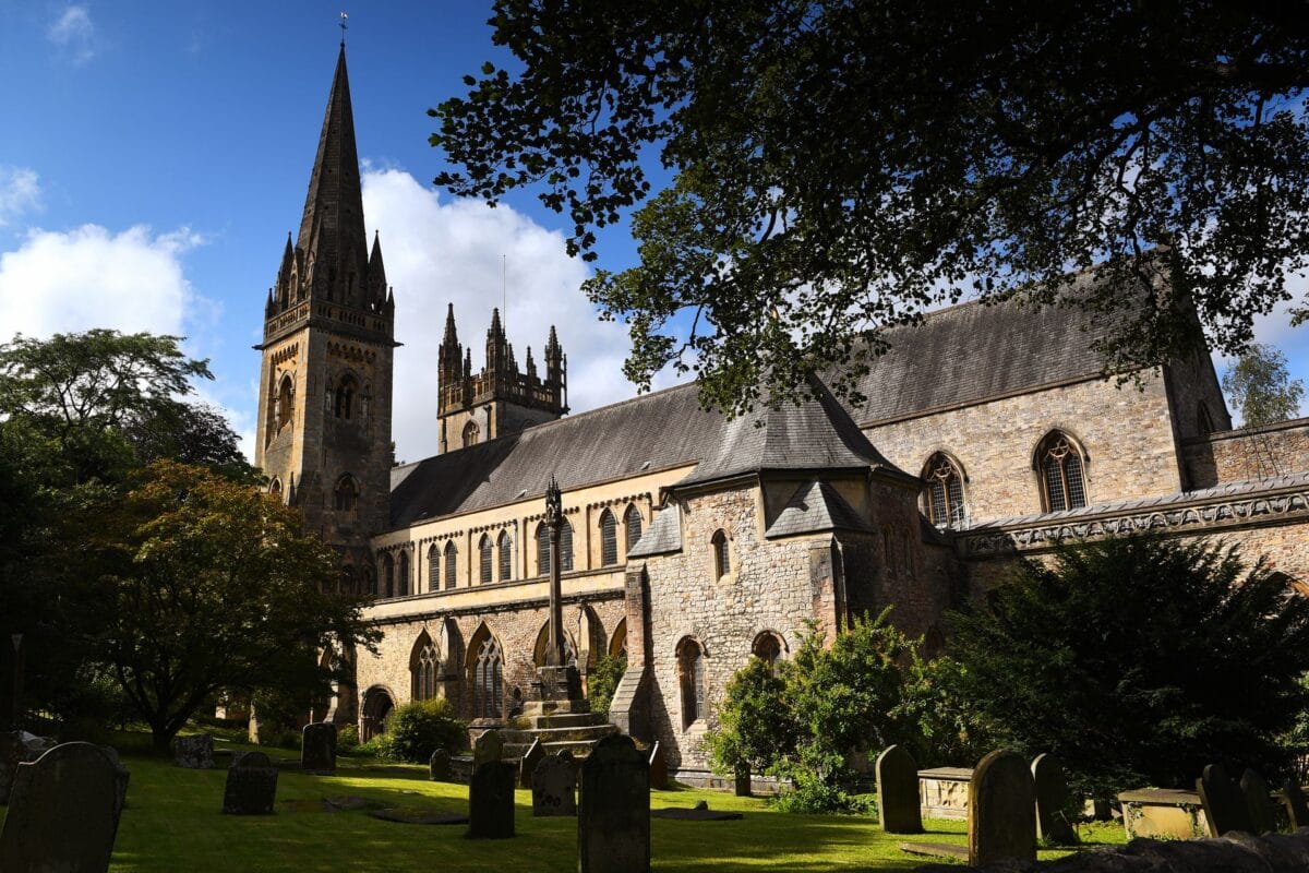 The stone exterior and tall, pointed spire of Llandaff Cathedral in Cardiff, rising above a peaceful green churchyard filled with old headstones on a bright, sunny day.