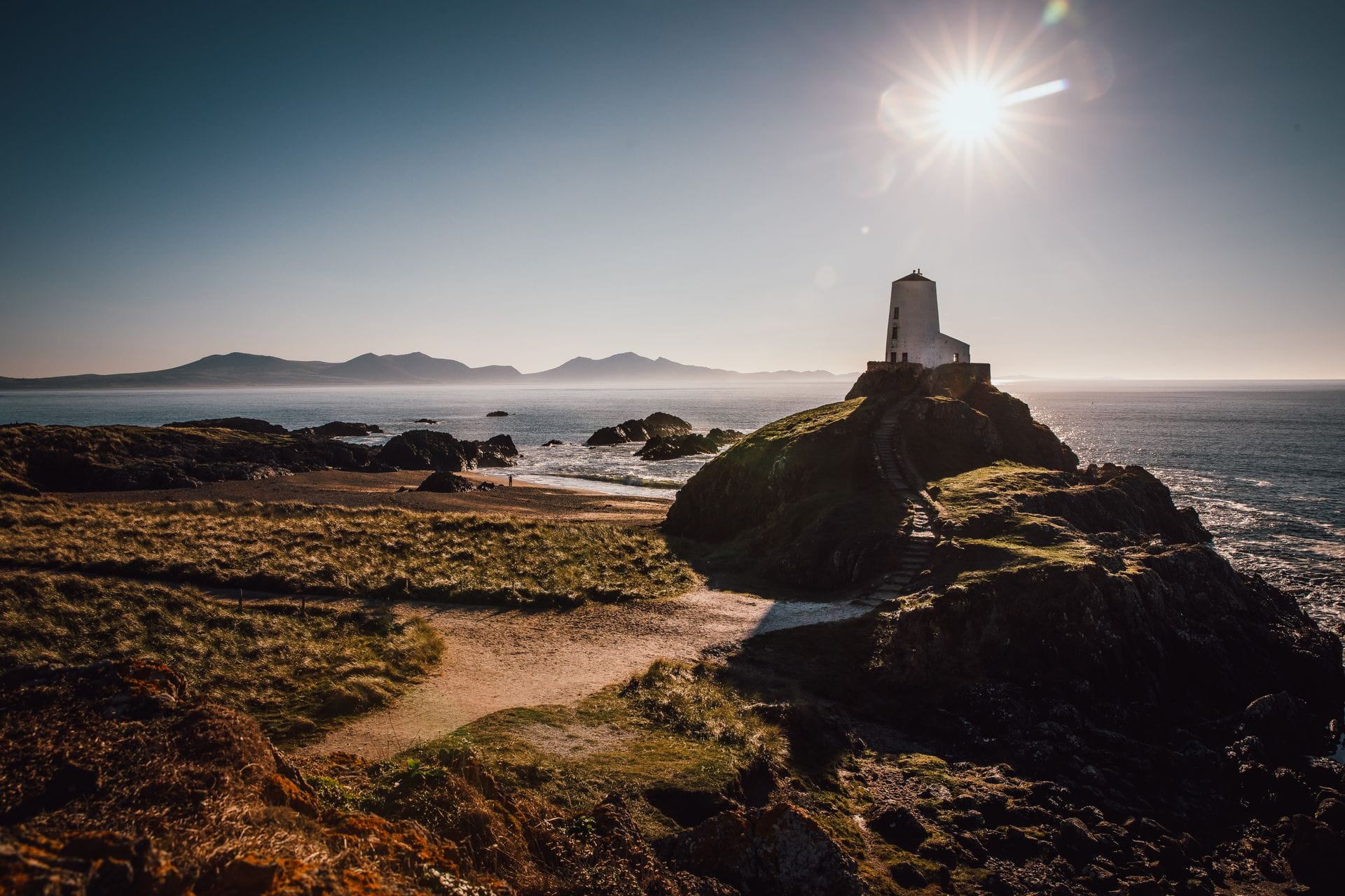 The Celtic cross and ruined chapel of St Dwynwen on Ynys Llanddwyn, a small tidal island off the coast of Anglesey, with the Eryri mountains visible across the sea on a clear January afternoon.