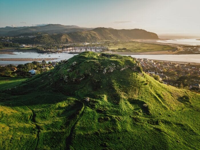 A lush green hill rises above a scenic landscape near Colwyn Bay, with a river, village houses, and distant mountains under a clear sunset sky. Paths wind through the hillside as soft sunlight highlights the vibrant greenery.