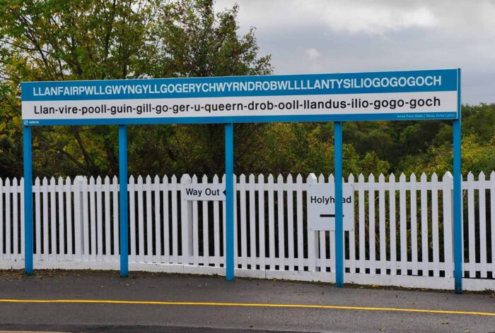 A railway station sign displays the world's longest Welsh place name Llanfairpwllgwyngyllgogerychwyrndrobwllllantysiliogogogoch—an iconic stop for anyone exploring things to do in Wales. A white fence and green trees are visible in the background.