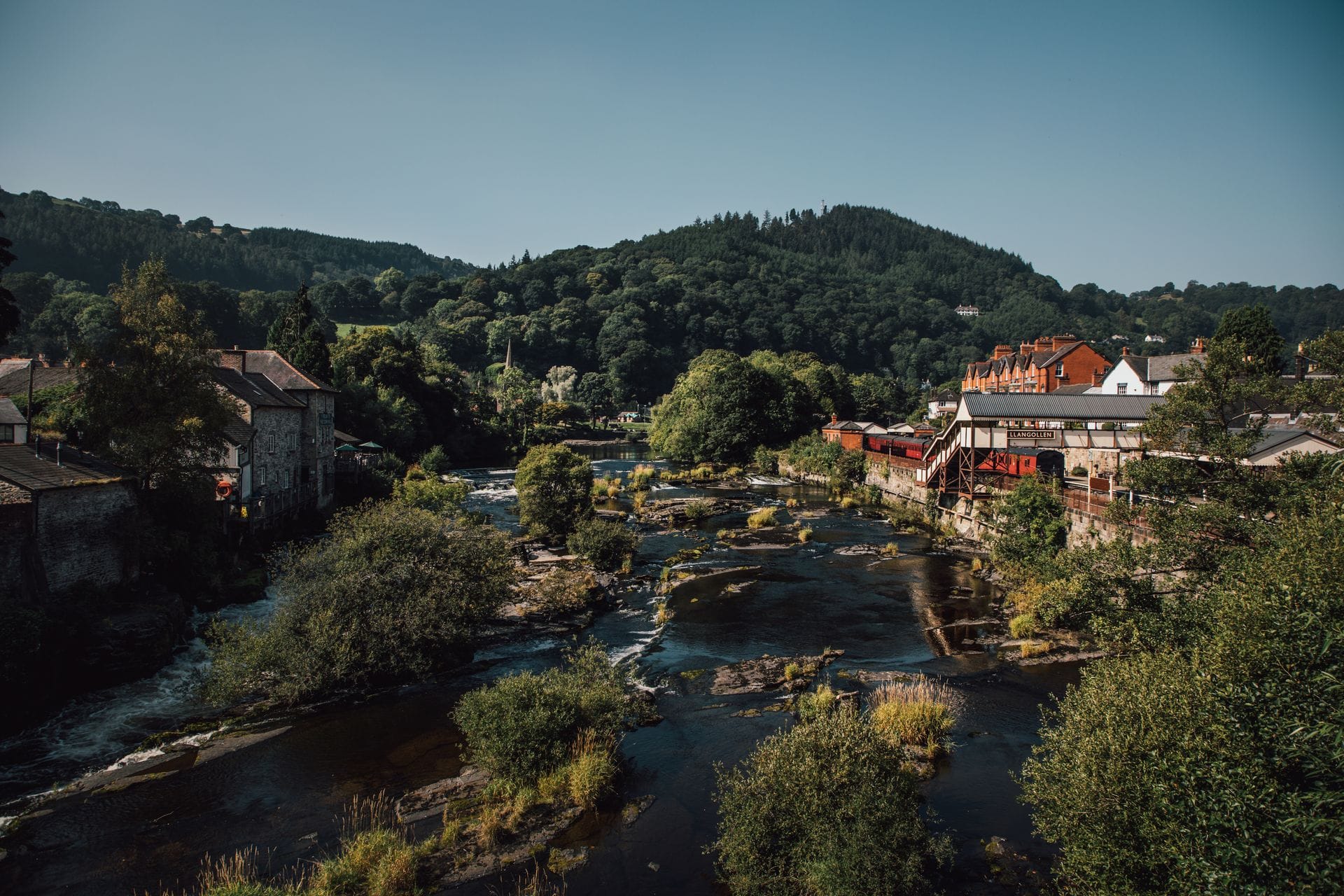 A scenic view of the River Dee flowing over a rocky bed through the picturesque town of Llangollen. Historic stone buildings and riverside businesses line the lush green banks, nestled beneath large, heavily forested rolling hills under a clear blue sky.