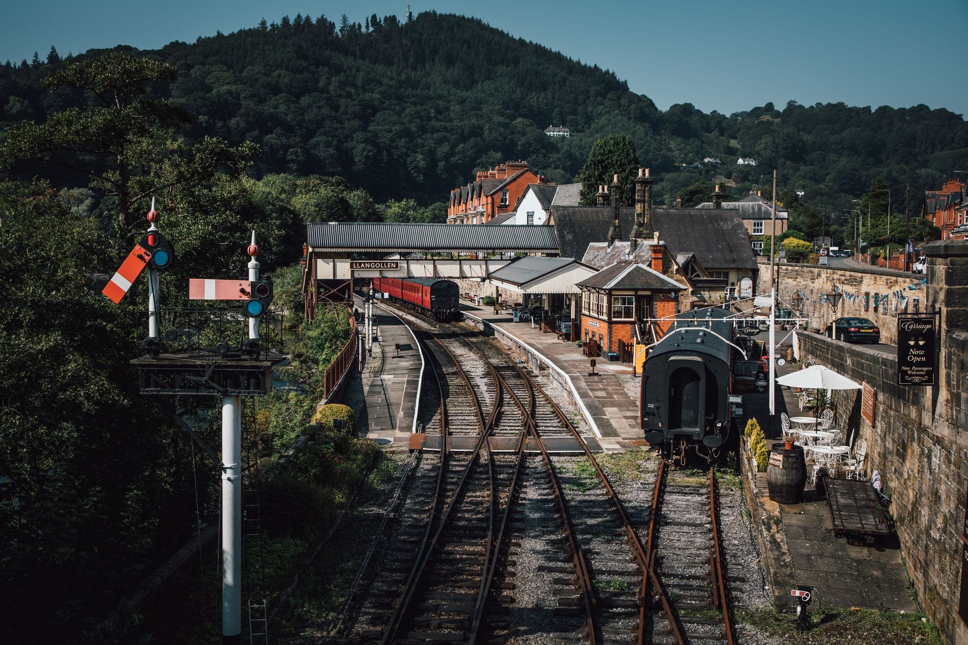 A scenic view of the historic Llangollen Railway station. The image features curving railway tracks leading towards a pedestrian footbridge and platforms with vintage train carriages. Classic semaphore signals stand in the foreground, set against a picturesque backdrop of charming town buildings and a steep, densely forested hill under a bright blue sky.