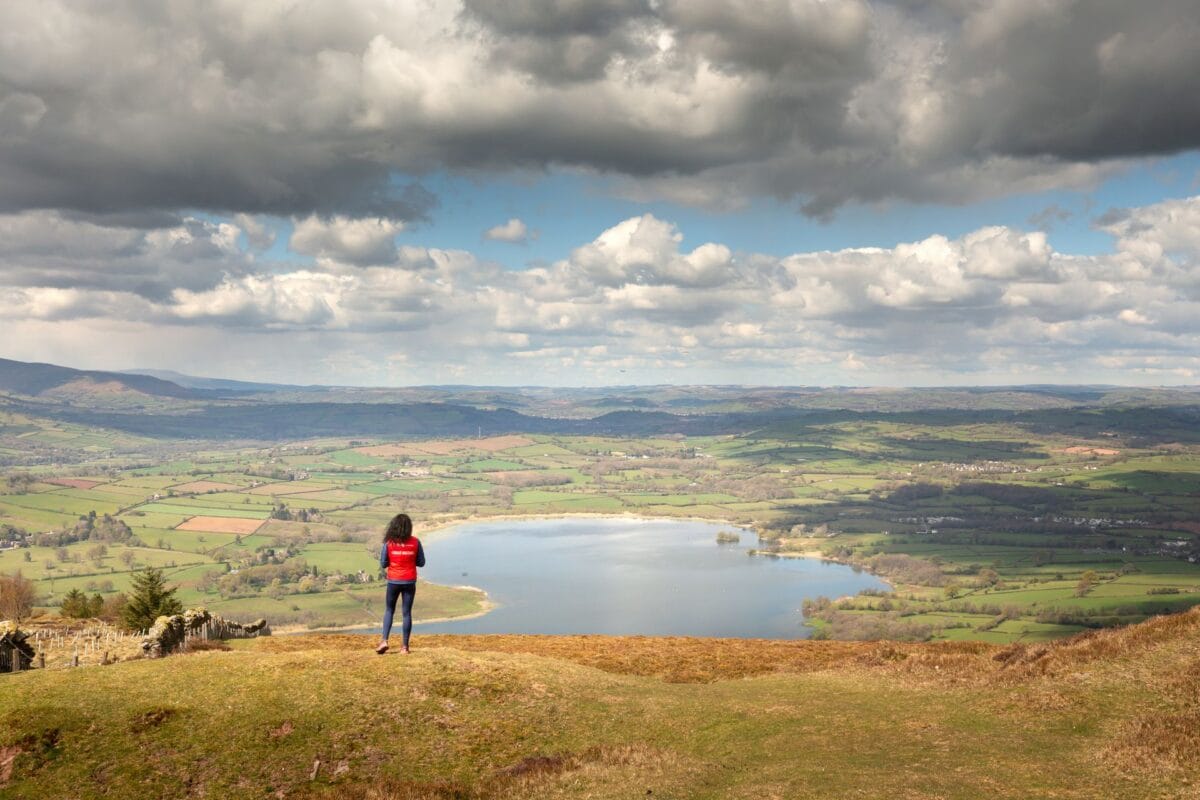A person standing on a grassy ridge looks out over the wide, blue waters of Llangorse Lake in the Brecon Beacons, surrounded by a patchwork of green farm fields and distant hills under a heavy, dramatic sky.