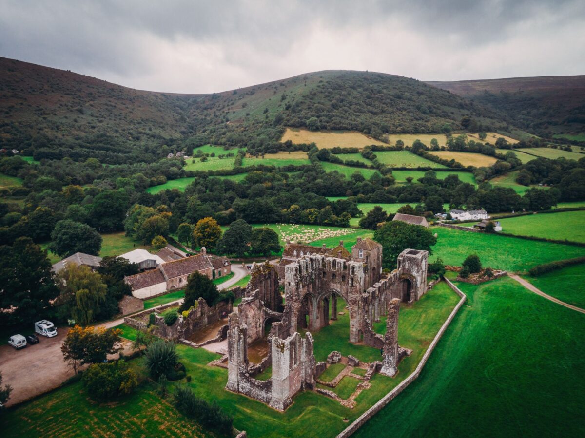 Elevated aerial view of the Llanthony Priory ruins. The roofless, historic stone arches and walls sit on bright green grass alongside a small farm and car park, surrounded by the steep, rolling hills and patchwork fields of the Offa's Dyke hiking route.