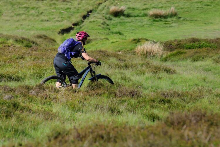 A person wearing a purple helmet and rucksack rides a mountain bike through a grassy, green landscape with patches of bushes and uneven terrain. Enjoying the cycling and mountain biking in Wales