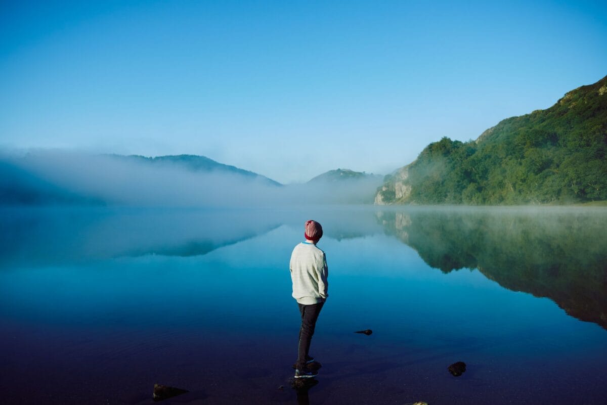 A person standing at the edge of the perfectly still, bright blue waters of Llyn Gwynant, looking out towards the surrounding green mountains partially obscured by a thick layer of morning mist.