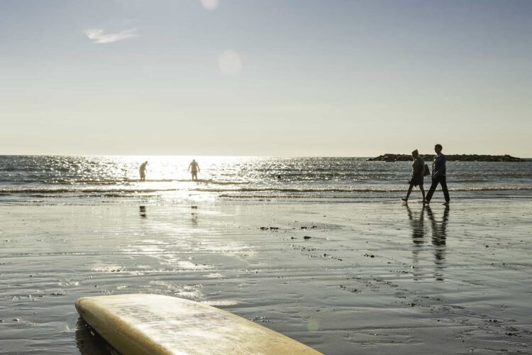 A yellow surfboard lies on wet sand in the foreground, whilst two people walk along the shoreline and two more stand in the shallow water—an idyllic scene near campsites near the beach in Wales, with sunlight glinting off the sea.