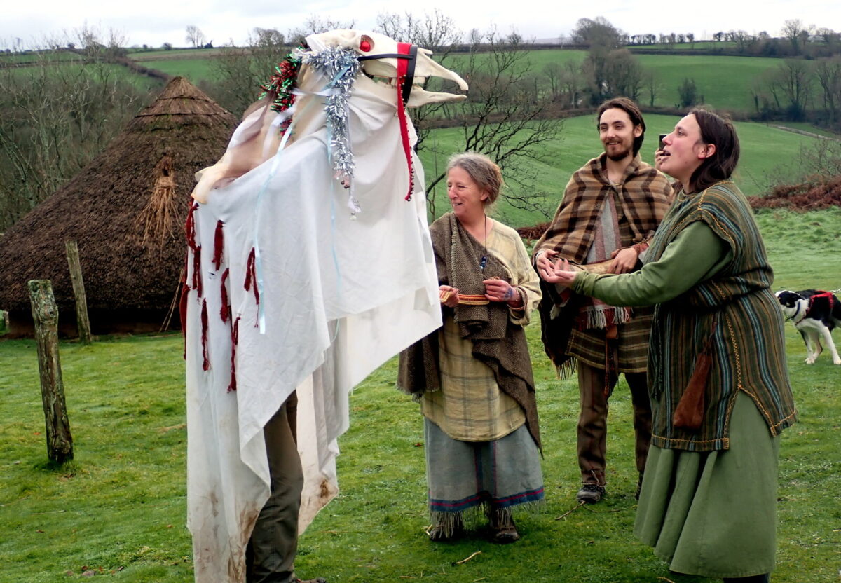 A traditional Mari Lwyd horse skull effigy draped in a white sheet and decorated with ribbons, being paraded outdoors in a grassy field. Three people wearing rustic, historical clothing stand before it, with one woman in a green tunic singing or speaking animatedly towards the effigy. A thatched roundhouse and a dog are visible in the background.