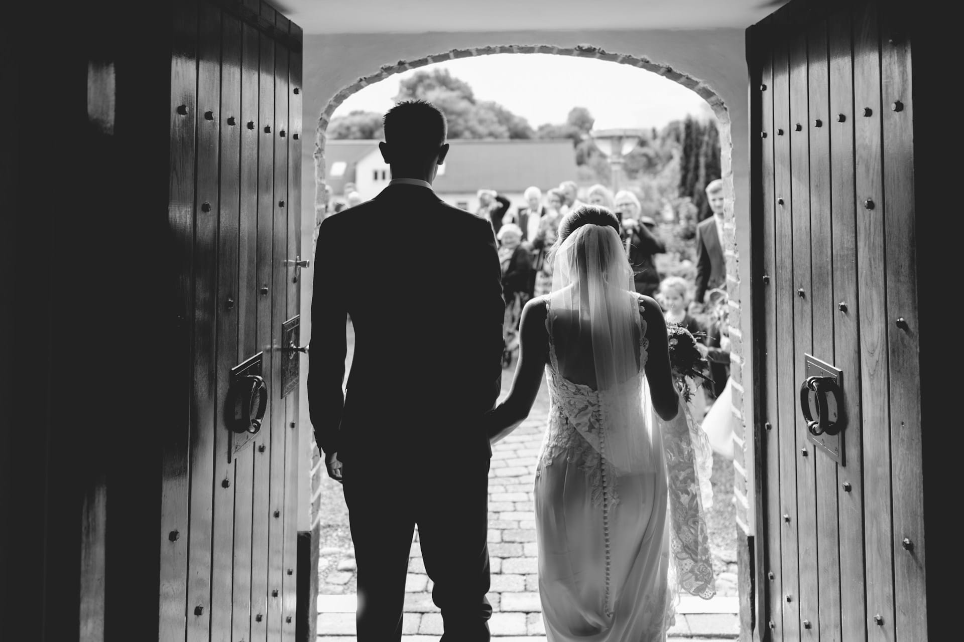 A black-and-white photograph taken from behind a bride and groom holding hands. They are silhouetted as they walk through large, open double wooden doors out into bright daylight toward a crowd of waiting guests.