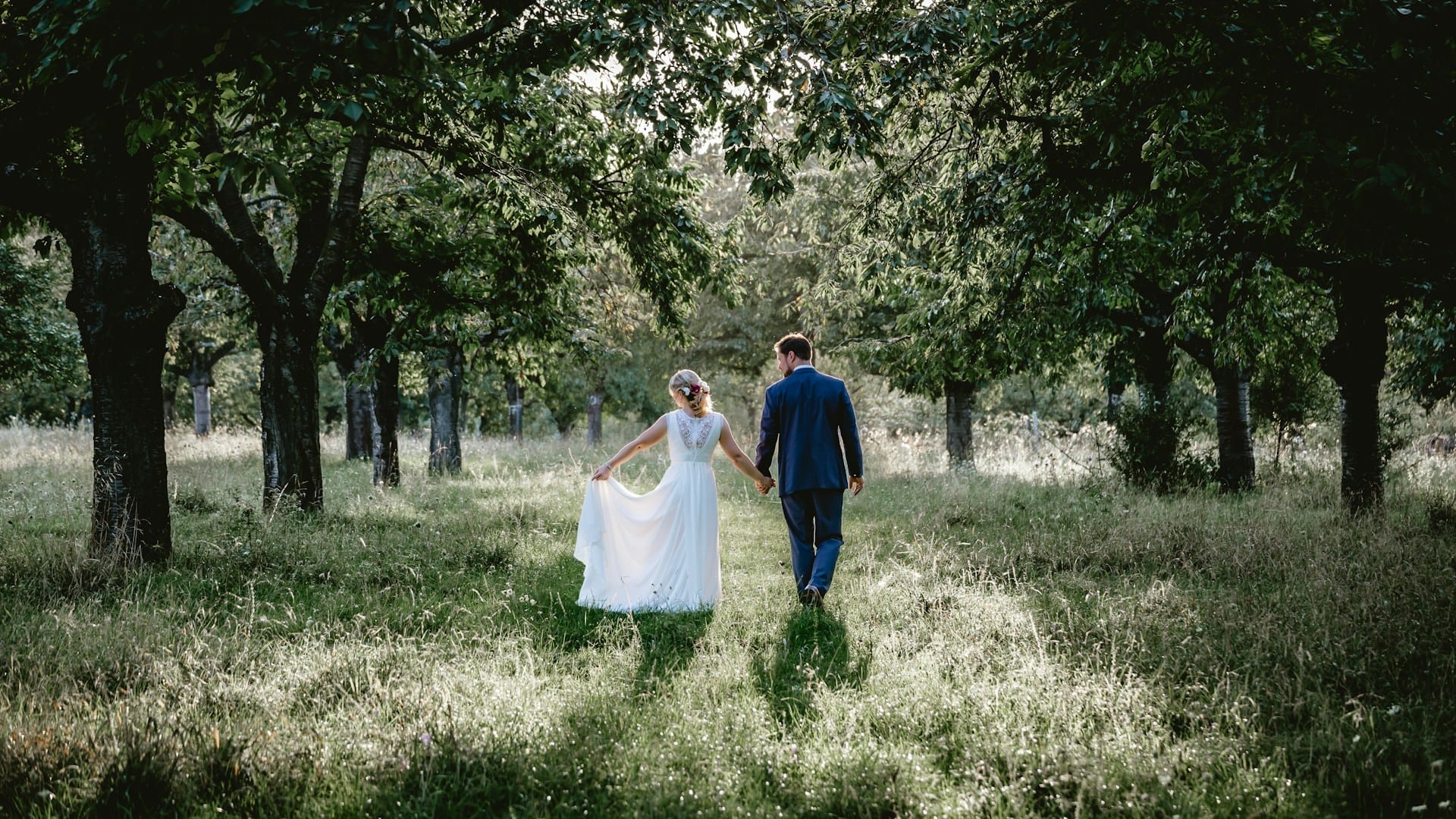 A bride and groom holding hands as they walk away from the camera through a sun-dappled orchard. The bride is gently lifting the skirt of her flowing white dress as they stroll through the long grass beneath a canopy of mature trees