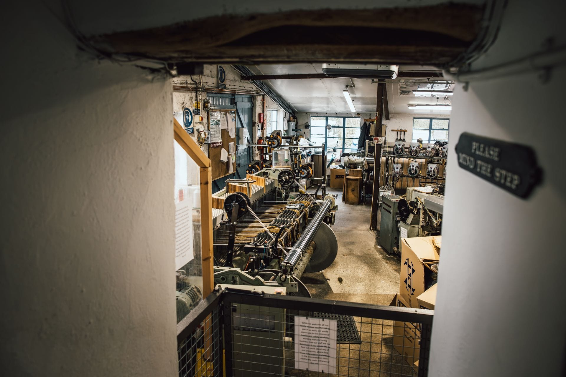 A working shuttle loom mid-weave inside a traditional Welsh woollen mill, seen through an open doorway, with warp threads stretched across the machine and cardboard boxes of finished stock stacked nearby.